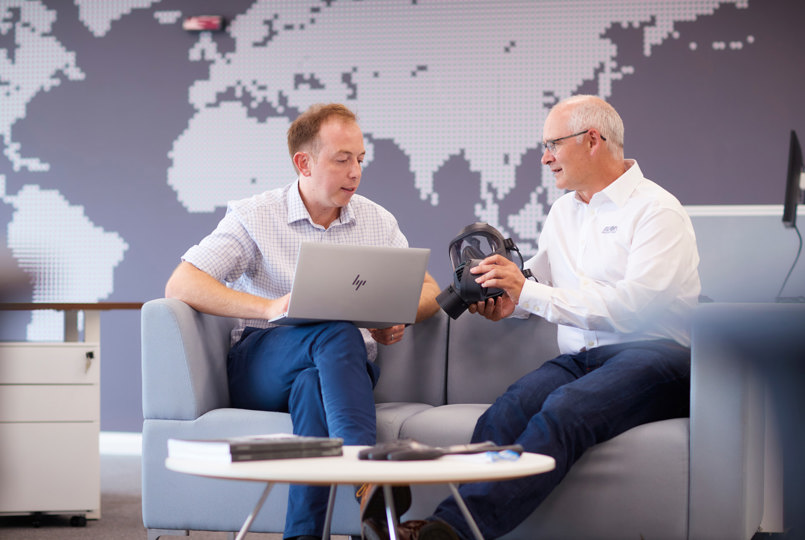 Two employees sat in an office looking at a respirator and laptop